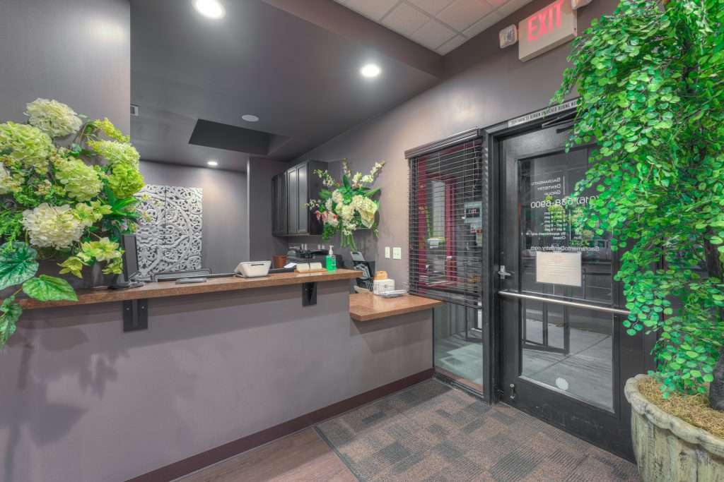 Front desk and entrance at Sacramento Dentistry Group with a glass door, fresh flowers, and a welcoming greenery display
